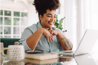 Uma mulher sentada em uma cadeira e com os braços apoiados em uma mesa está sorrindo olhando para o computador que está sobre a mesa